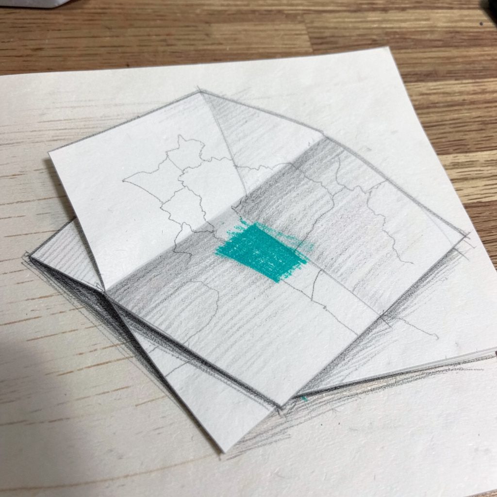 Graphite pencil sketch of a folded paper map resting on a wooden surface, light grey tones